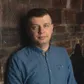 A man in a blue button-up shirt stands against a textured brick wall, looking at the camera with a neutral expression.