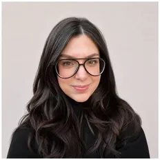 Person with long dark hair and large glasses, wearing a black turtleneck, smiles softly against a light background.