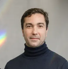 A man in a dark turtleneck sweater poses with a neutral expression against a light background with a hint of rainbow light.
