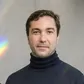 A man in a dark turtleneck sweater poses with a neutral expression against a light background with a hint of rainbow light.