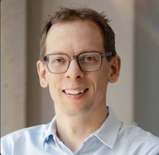 A man with glasses and short hair smiling, wearing a light blue shirt, standing in a well-lit room.