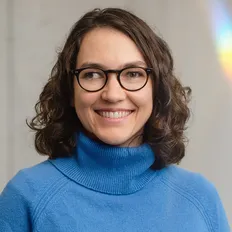 Smiling woman with curly hair and glasses, wearing a blue sweater, standing against a light background.