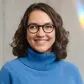 Smiling woman with curly hair and glasses, wearing a blue sweater, standing against a light background.