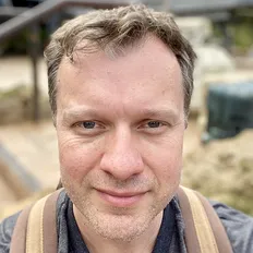 A man with short hair and a slight smile, wearing a backpack, outdoors with blurred background.