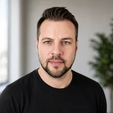 Man with short hair and beard, wearing a black shirt, looking at the camera in a bright room.