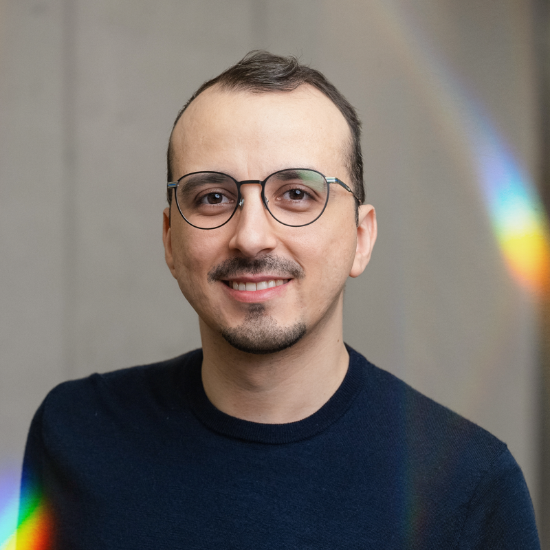 Smiling person with glasses and short hair, wearing a dark shirt. Light rainbow reflections adorn the image.