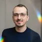Smiling person with glasses and short hair, wearing a dark shirt. Light rainbow reflections adorn the image.