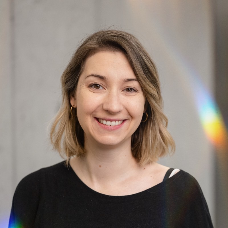 Smiling woman with shoulder-length hair and hoop earrings, wearing a black top against a neutral background with faint rainbow reflections.