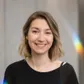 Smiling woman with shoulder-length hair and hoop earrings, wearing a black top against a neutral background with faint rainbow reflections.