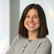 Smiling woman with shoulder-length brown hair, wearing a gray sweater, standing against a plain background.