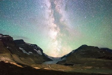Milky Way core rising above a glacial mountain valley under a green-tinted, star-filled night sky.