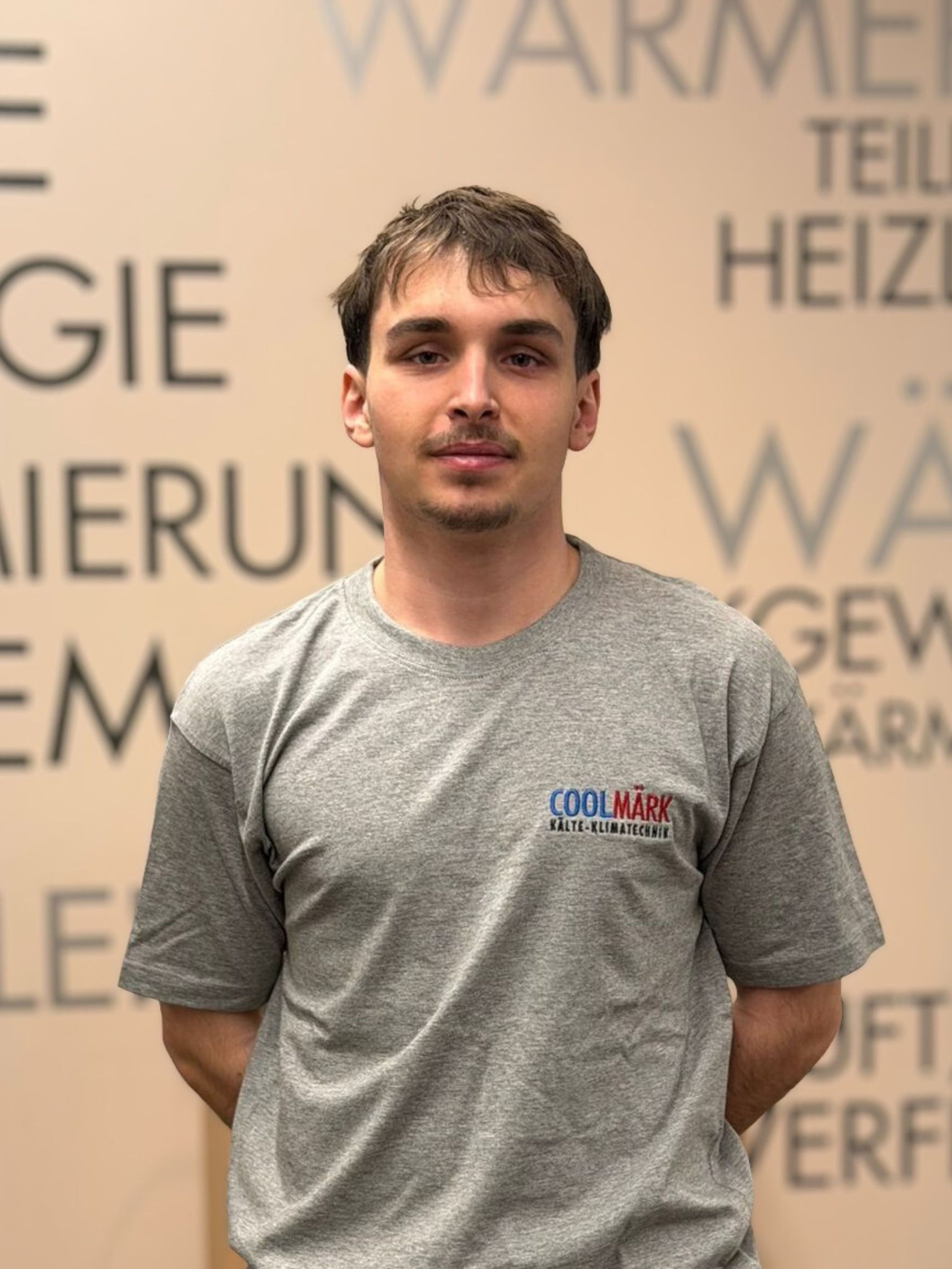 Young man with short hair in a gray Cool Mark shirt stands against a wall with partial German text in the background.