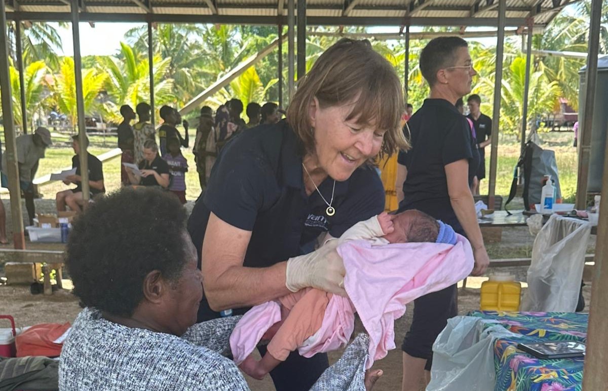 A woman, Catharina, smiling while handing a newborn wrapped in a pink blanket to another person, with an outdoor gathering in the background.