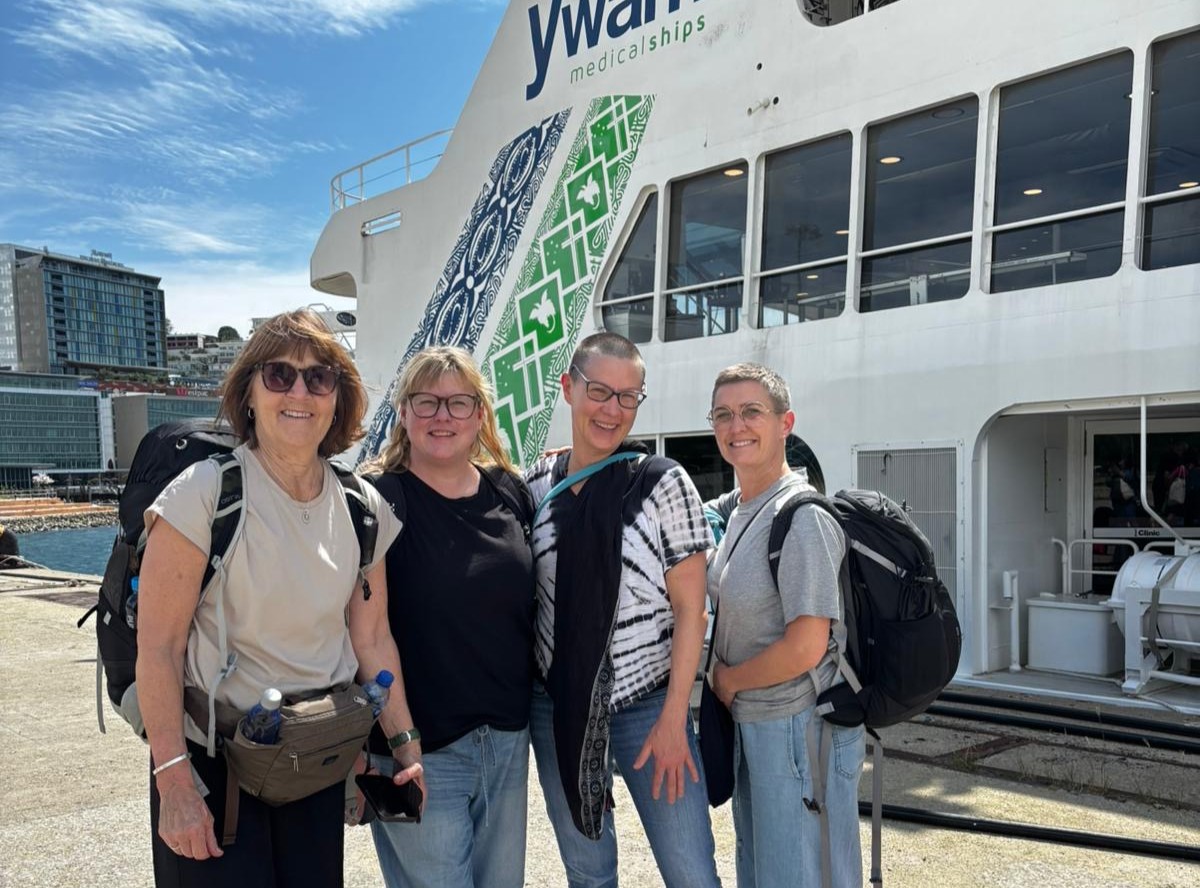 Four women with backpacks stand smiling in front of a ship at a dock, with buildings in the background.