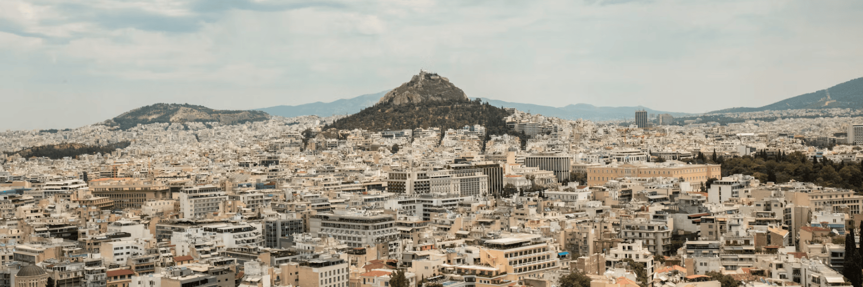Panoramic view of Athens with densely packed buildings and a hill in the background under a cloudy sky.
