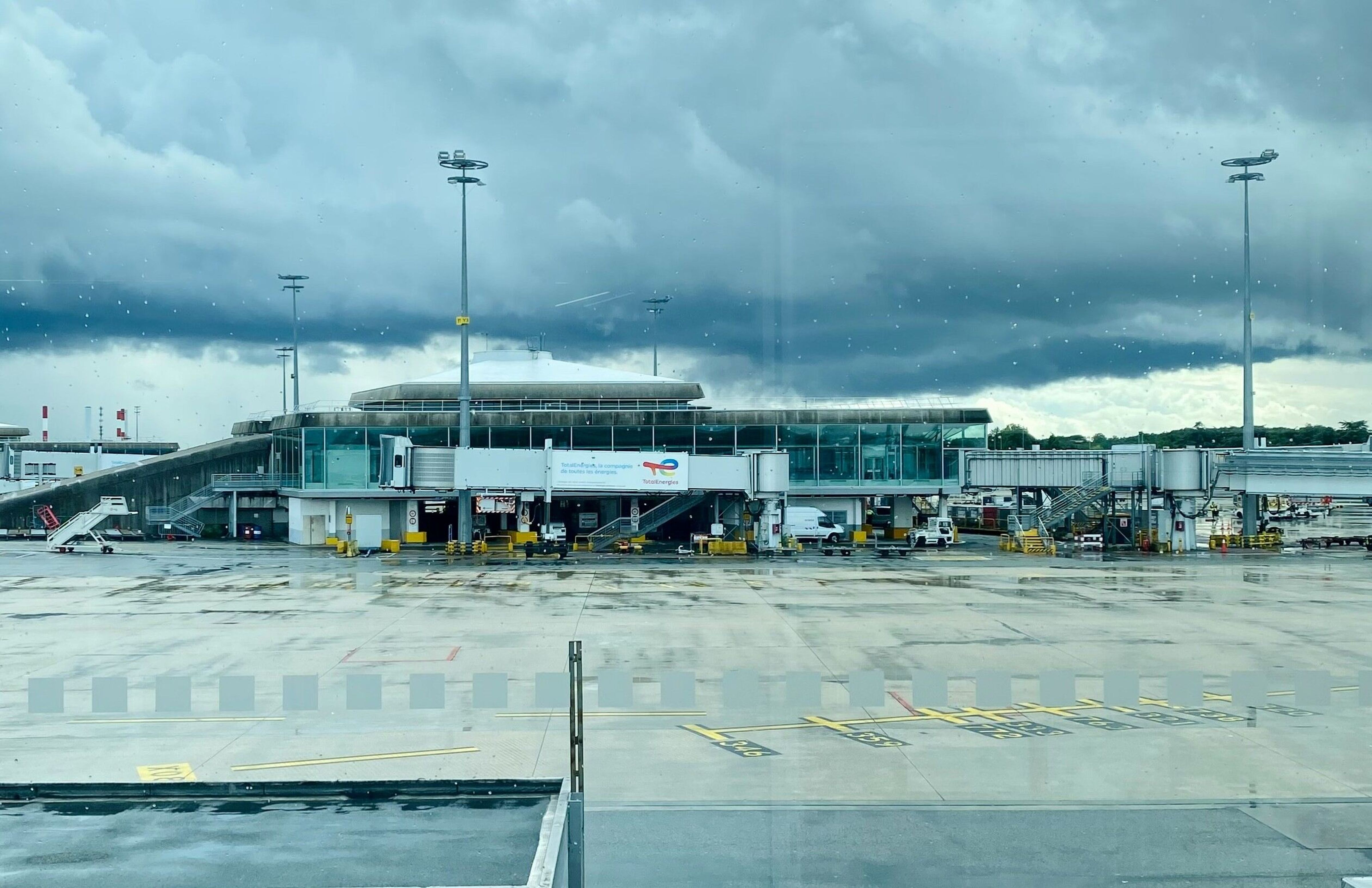 Empty airport tarmac with a terminal building under a cloudy sky, featuring rain and distant vehicles.