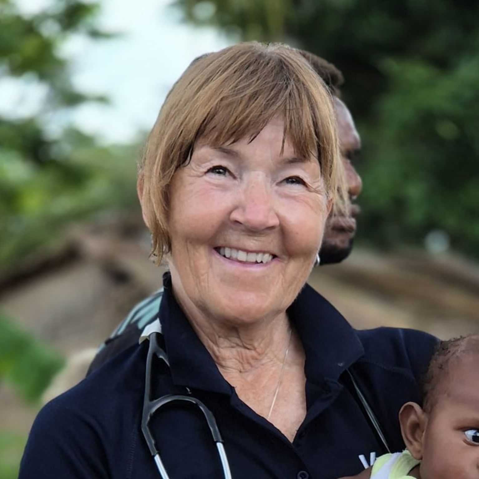 Smiling woman outdoors with a stethoscope around her neck, holding a small child. Green foliage in the background.