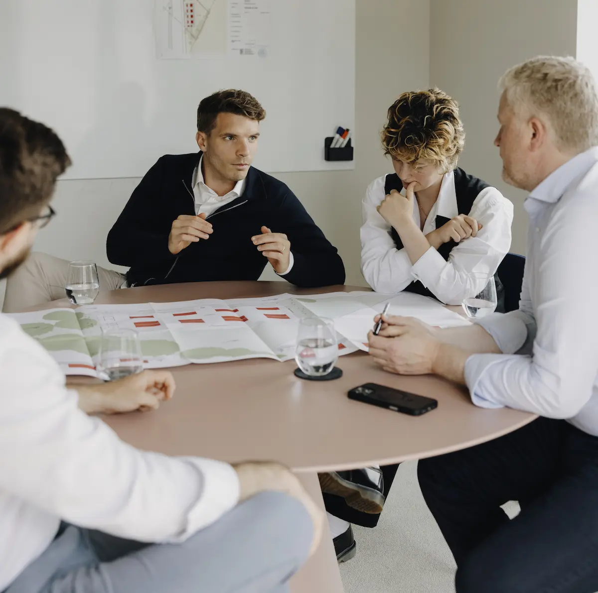 Four people sit around a table discussing architectural plans, with diagrams and glasses of water visible.