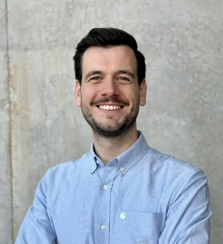 A smiling man with dark hair and a beard stands in front of a gray textured wall, wearing a light blue button-up shirt.