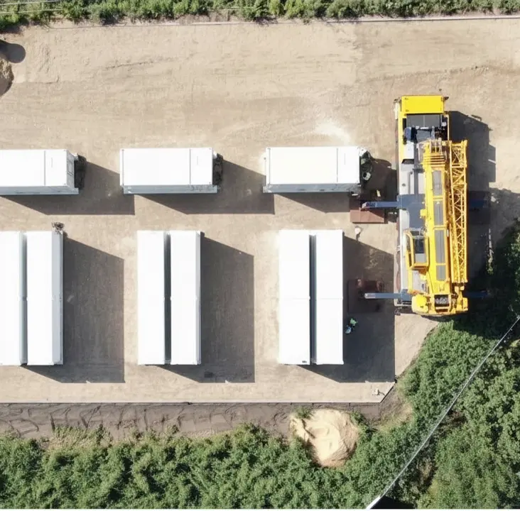 Aerial view of six white rectangular storage containers and a yellow construction vehicle on a dirt lot, surrounded by greenery.