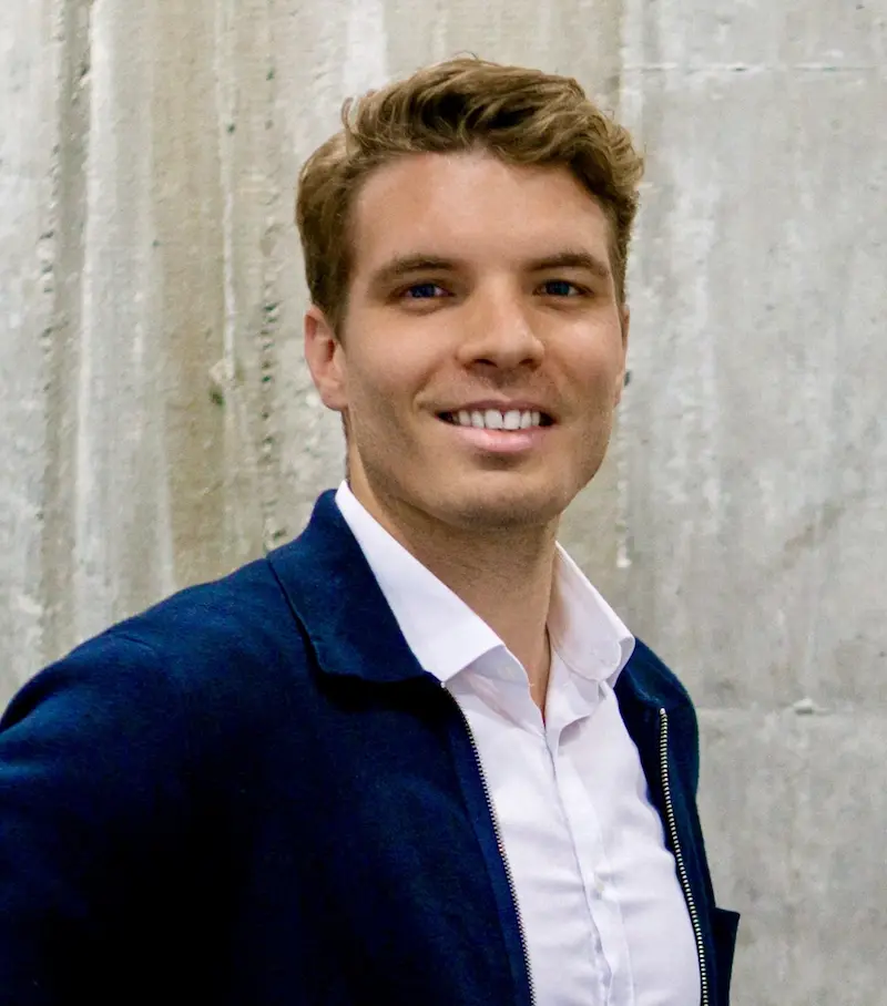 Smiling person with short hair wearing a navy blazer and white shirt, standing in front of a textured concrete wall.
