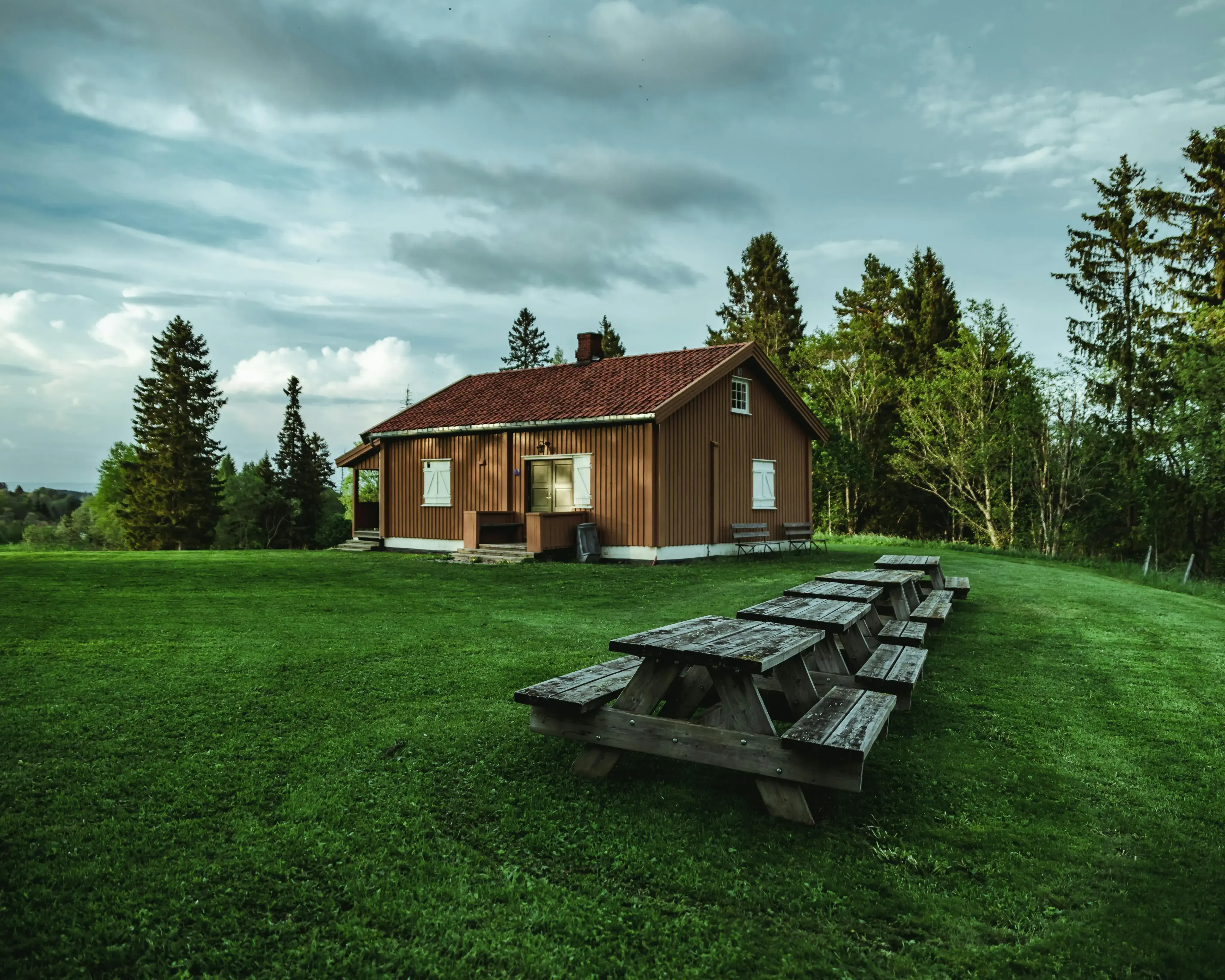A rustic wooden cabin with a red roof sits on a lush green lawn, surrounded by trees, with picnic tables in the foreground under a cloudy sky.