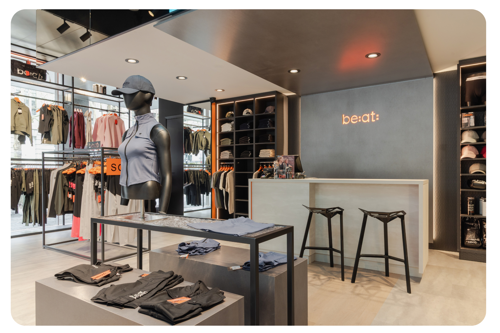 Interior of a modern clothing store with a mannequin, folded apparel, display racks, hats on shelves, and a counter with stools.