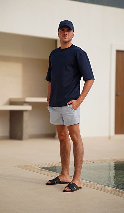 Man in a navy shirt, gray shorts, and black sandals stands by a small pool, hands in pockets, with a modern building in the background.