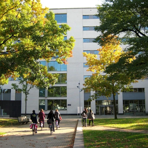 Campus at Tilburg University. People biking and walking on a path lined with trees, leading to a modern building with large windows on a sunny day.