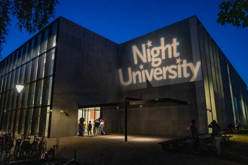 Building exterior at night with "Night University" projected on the wall, people entering, and a dark blue sky.