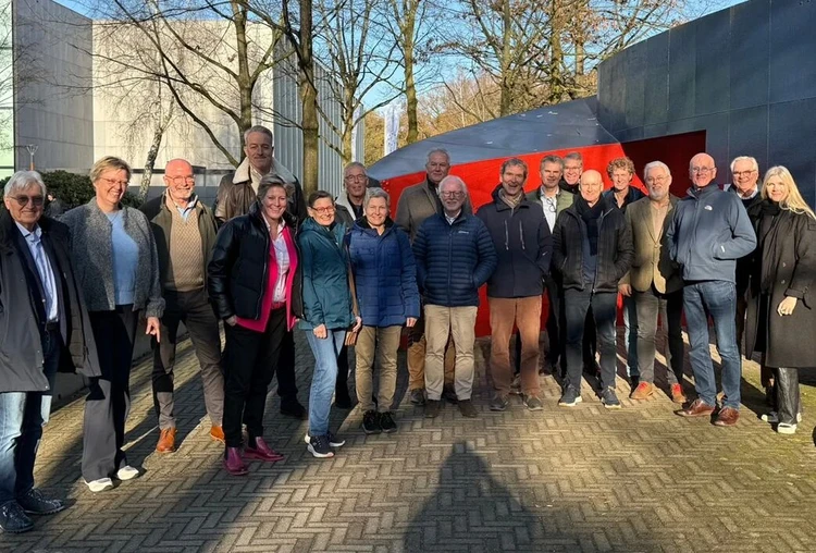 A group of people stand smiling outdoors, posing together on a paved area with trees and buildings in the background.