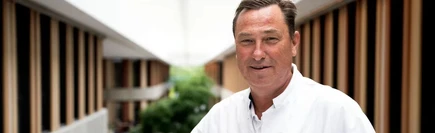 A man in a white shirt stands in a modern building with wooden accents and large windows.