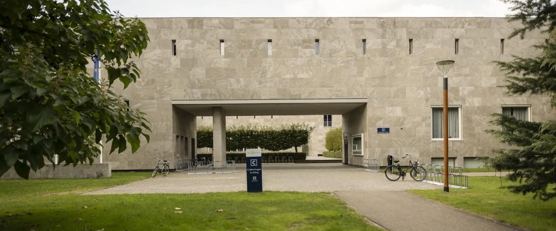 Modern building with rectangular openings, surrounded by greenery, bicycles parked nearby.