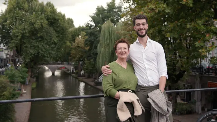 A smiling man and woman standing on a canal bridge with lush greenery and overcast sky in the background.