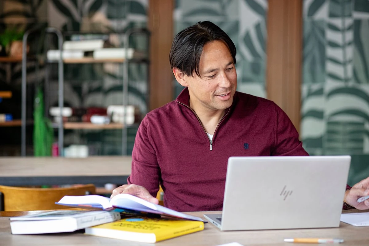 Man in a maroon sweater works on a laptop at a table with open books, in a room with leafy wallpaper and shelves in the background.