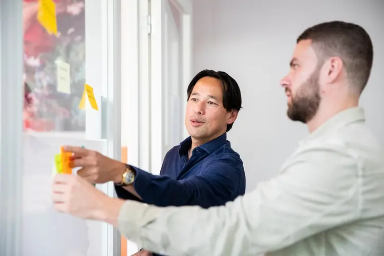 Two men collaborate at a whiteboard, attaching sticky notes. One points while discussing ideas in a bright, modern office setting.