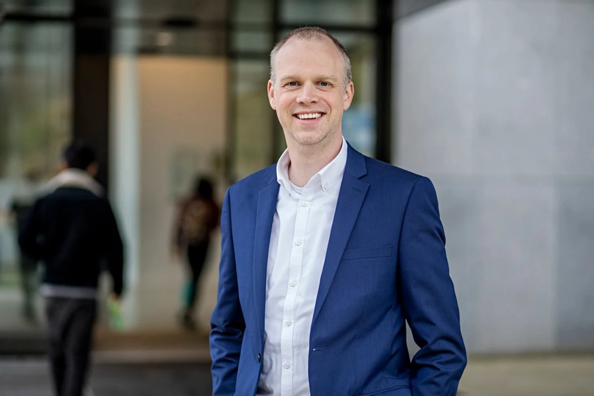 Smiling man in a blue suit and white shirt stands outside a building entrance, with people walking in the background.