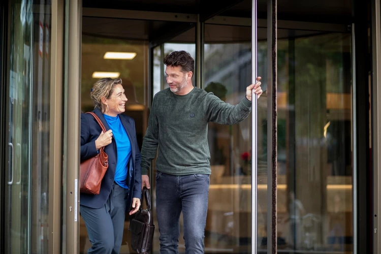 Man holding door open for woman as they exit a building. Both are smiling and carrying bags. Glass doors and interior visible in the background.
