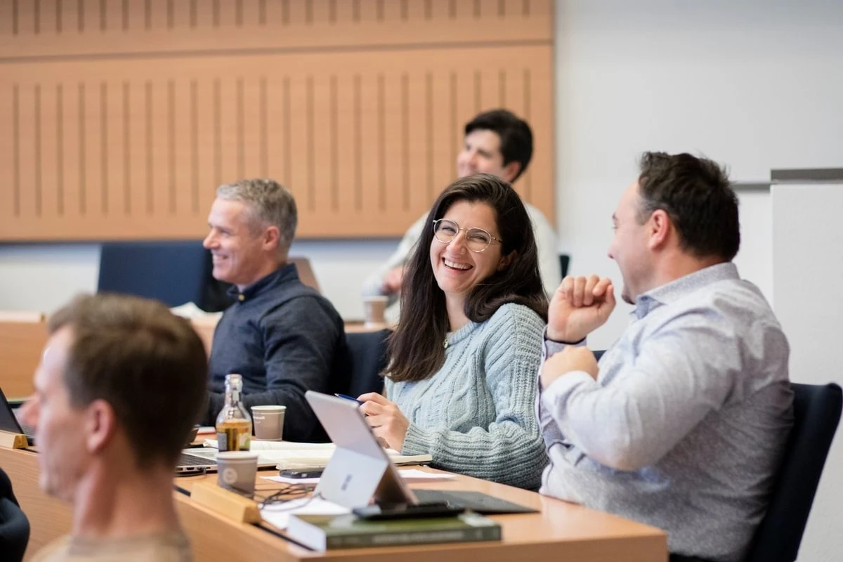 People seated in a classroom, smiling and engaging in conversation, with notebooks and laptops on the desks.