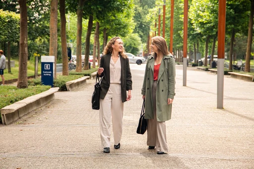 Two women walking and talking on a tree-lined path, both carrying bags. One wears a beige suit, the other a green coat over a red top.