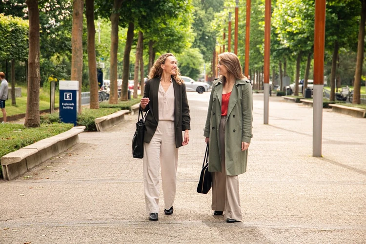 Two women walking and talking on a tree-lined path, both carrying bags. One wears a beige suit, the other a green coat over a red top.