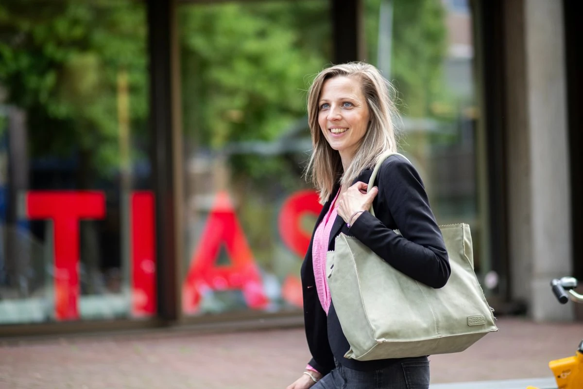 Smiling woman with a beige bag walks outside a building with large red letters on the glass.