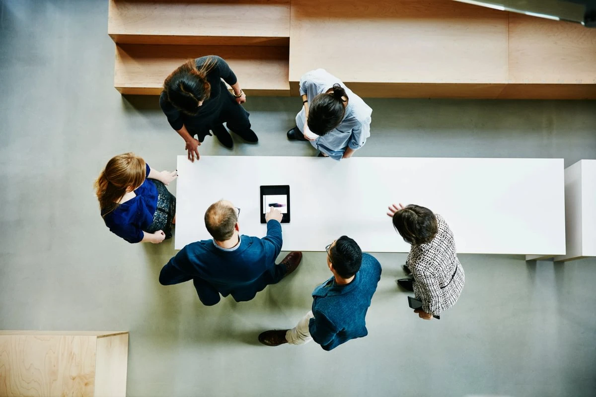 Aerial view of six people gathered around a white table, discussing and pointing at a tablet.