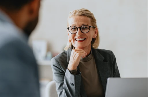 Smiling woman in glasses and a blazer sits at a desk, engaging in conversation with a blurred person in the foreground.
