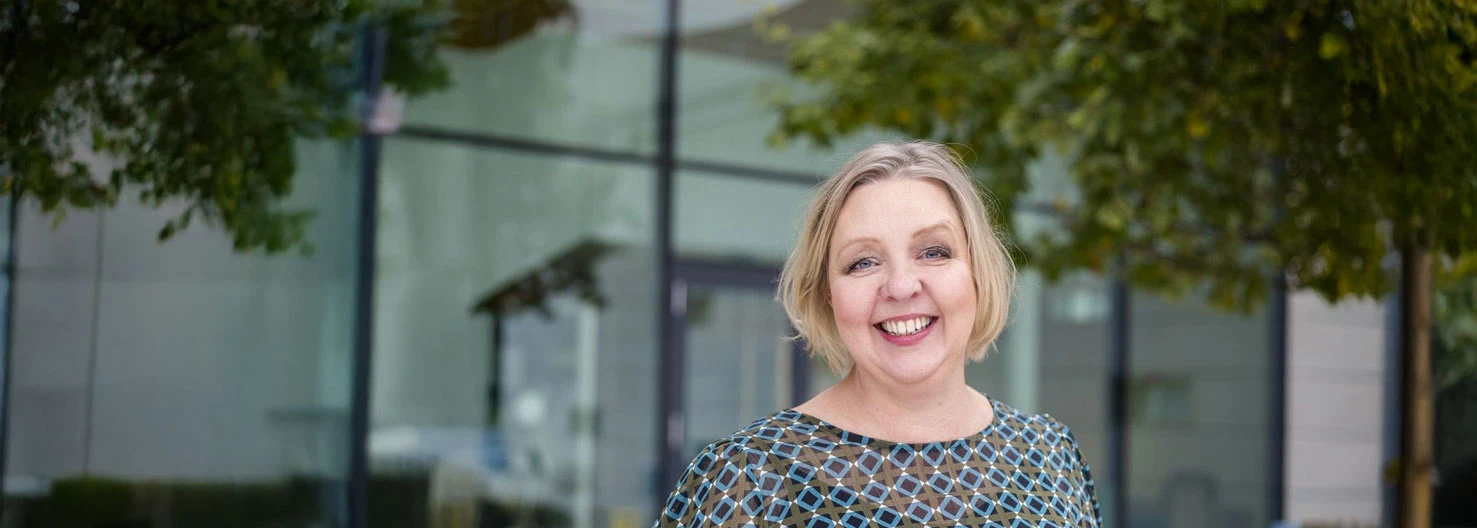 Smiling woman standing outdoors in front of a modern glass building with trees.