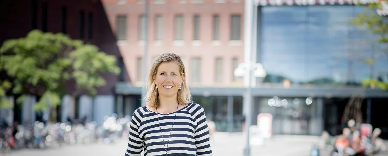 Woman smiling outdoors, standing in front of a modern building with large windows and trees in the background.