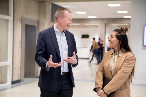 A man in a suit and a woman in a beige blazer are talking in a hallway, with people and doors in the background.