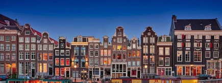Row of traditional Dutch canal houses at dusk, with warm lights glowing from windows, under a clear blue sky.