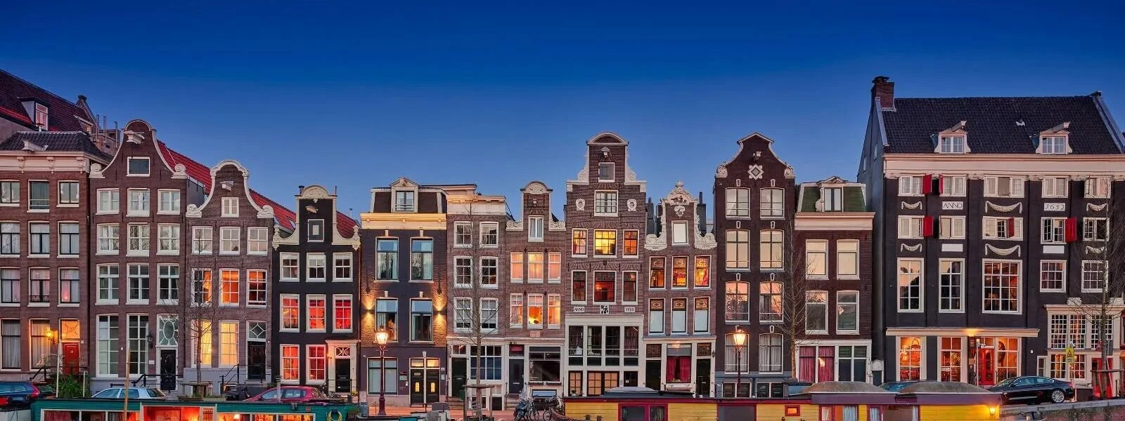 Row of traditional Dutch canal houses at dusk, with warm lights glowing from windows, under a clear blue sky.