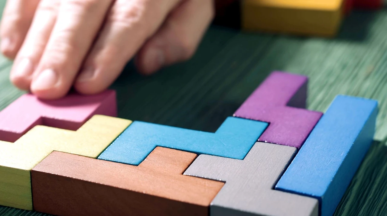Close-up of a hand arranging colorful, interlocking wooden blocks on a green surface, resembling a puzzle.